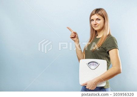 Woman holding scale to balance weight loss pointing with finger to empty space Woman holding scale to balance weight loss pointing with finger to empty space 103956320