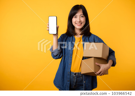 Portrait asian young woman wearing yellow t-shirt and denim shirt holding mobile phone and parcel box isolated on yellow studio background, Delivery courier and shipping service concept. Portrait asian young woman wearing yellow t-shirt and denim shirt holding mobile phone and parcel box isolated on yellow studio background, Delivery courier and shipping service concept. 103966542