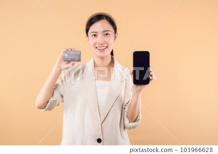portrait of happy successful confident young asian business woman wearing white jacket holding smartphone and credit card standing over beige background. shopping concept. 103966628