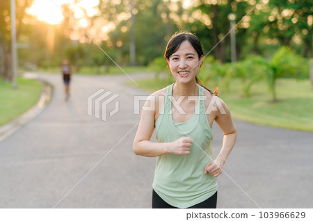 Fit Asian young woman jogging in park smiling happy running and enjoying a healthy outdoor lifestyle. Female jogger. Fitness runner girl in public park. healthy lifestyle and wellness being concept 103966629