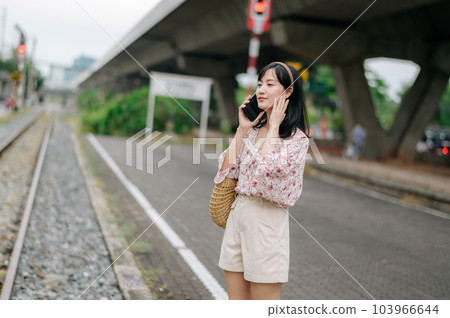Asian young woman traveler with weaving basket using a mobile phone beside railway train station in Bangkok. Journey trip lifestyle, world travel explorer or Asia summer tourism concept. 103966644