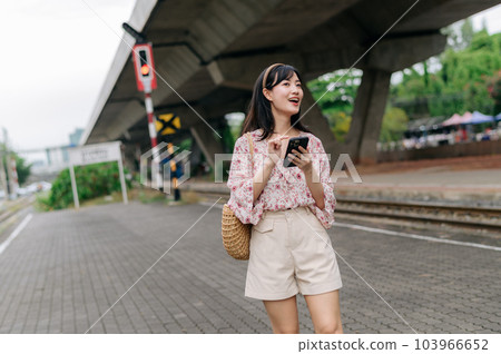 Asian young woman traveler with weaving basket using a mobile phone beside railway train station in Bangkok. Journey trip lifestyle, world travel explorer or Asia summer tourism concept. 103966652