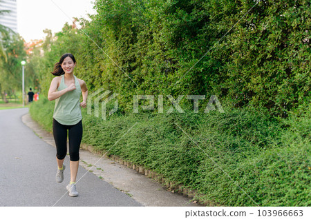 Fit Asian young woman jogging in park smiling happy running and enjoying a healthy outdoor lifestyle. Female jogger. Fitness runner girl in public park. healthy lifestyle and wellness being concept 103966663