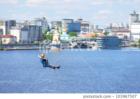 A tourist is speeding down a zipline against the backdrop of a blurred river and city. A tourist is speeding down a zipline against the backdrop of a blurred river and city. 103967507