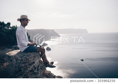 Digital nomad, man in the hat, a businessman with a laptop sits on the rocks by the sea during sunset, makes a business transaction online from a distance. Remote work on vacation. 103968250