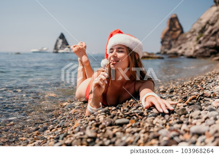 Woman travel sea. Happy tourist enjoy taking picture on the beach for memories. Woman traveler in Santa hat looks at camera on the sea bay, sharing travel adventure journey 103968264