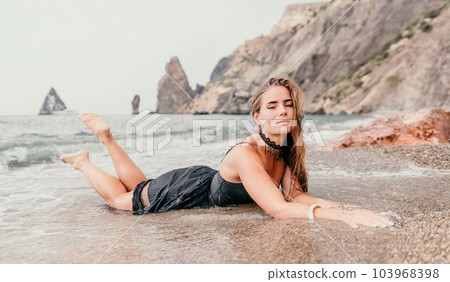 Woman summer travel sea. Happy tourist in black dress enjoy taking picture outdoors for memories. Woman traveler posing on sea beach surrounded by volcanic mountains, sharing travel adventure journey 103968398