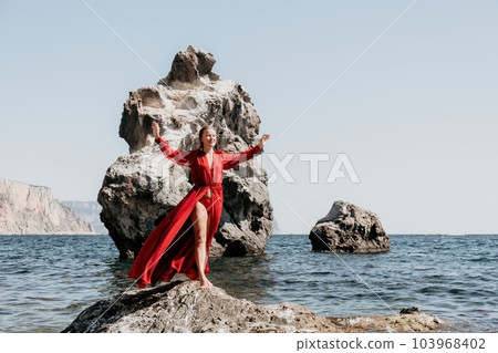 Woman travel sea. Happy tourist in long red dress enjoy taking picture outdoors for memories. Woman traveler posing on beach at sea surrounded by volcanic mountains, sharing travel adventure journey 103968402