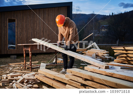 Man worker building wooden frame house. Carpenter using tape measure for measuring wooden planks and making marks with pencil. Carpentry concept. Man worker building wooden frame house. Carpenter using tape measure for measuring wooden planks and making marks with pencil. Carpentry concept. 103971818