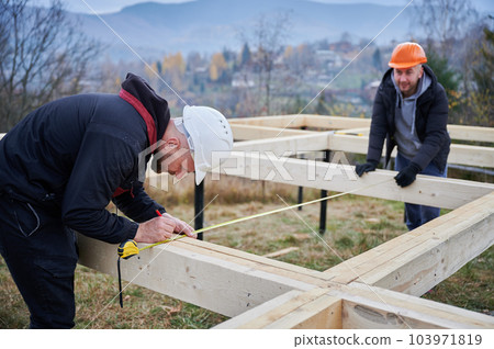 Man worker building wooden frame house on pile foundation. Carpenter using tape measure for measuring wooden planks and making marks with pencil. Carpentry concept. Man worker building wooden frame house on pile foundation. Carpenter using tape measure for measuring wooden planks and making marks with pencil. Carpentry concept. 103971819