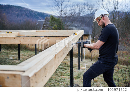 Man building wooden frame house on pile foundation. Male worker drilling hole by drill in timber framing of future house. Builder wearing white helmet and protective goggles. Carpentry concept. Man building wooden frame house on pile foundation. Male worker drilling hole by drill in timber framing of future house. Builder wearing white helmet and protective goggles. Carpentry concept. 103971822