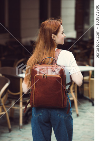 beautiful red-haired girl with red lips posing with a leather backpack. wearing blue jeans and a sweater beautiful red-haired girl with red lips posing with a leather backpack. wearing blue jeans and a sweater 103971900
