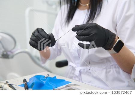 Close-up dental floss in the hands of a doctor dentist wearing black surgical gloves, working with gypsum dental model of human jaw in medical clinic. Dental health and oral hygiene concept. Dentistry Close-up dental floss in the hands of a doctor dentist wearing black surgical gloves, working with gypsum dental model of human jaw in medical clinic. Dental health and oral hygiene concept. Dentistry 103972467