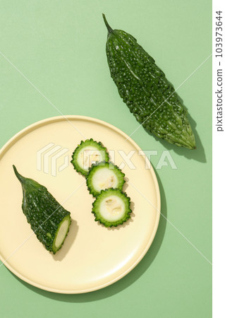 On the green background, a yellow dish with bitter melon cut in half and slices displayed on. Bitter melon (Momordica charantia) improves our immune system and respiratory health On the green background, a yellow dish with bitter melon cut in half and slices displayed on. Bitter melon (Momordica charantia) improves our immune system and respiratory health 103973644