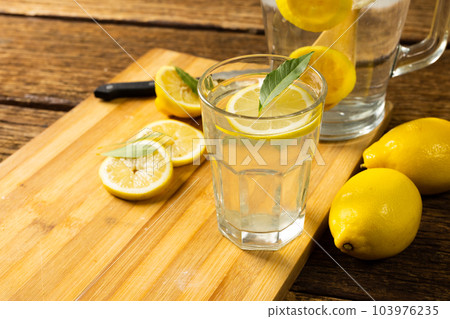 Close-up of lemonade in jug and drinking glass with lemons and leaves on cutting board over table 103976235