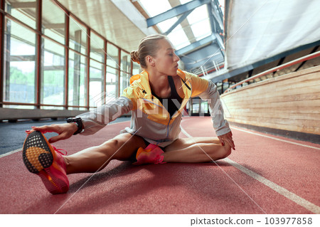 A young athlete girl is warming up before the race at the stadium. A slender blonde in a black tracksuit and a yellow-gray jacket stretches her muscles sitting on a cinder track. Sports and recreation A young athlete girl is warming up before the race at the stadium. A slender blonde in a black tracksuit and a yellow-gray jacket stretches her muscles sitting on a cinder track. Sports and recreation 103977858