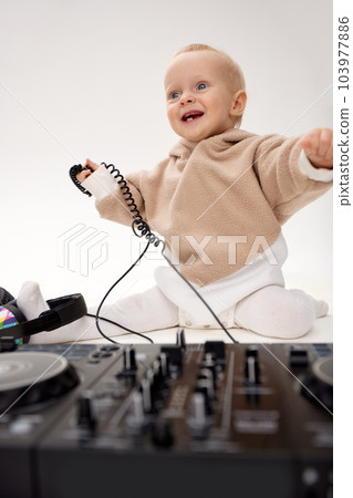 A cute smiling little child in a beige hoodie is sitting on the floor with dj headphones and a dj mixing board. Music and fun. Isolated on white background. A cute smiling little child in a beige hoodie is sitting on the floor with dj headphones and a dj mixing board. Music and fun. Isolated on white background. 103977886