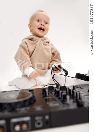 A cute smiling little child in a beige hoodie is sitting on the floor with dj headphones and a dj mixing board. Music and fun. Isolated on white background. 103977887