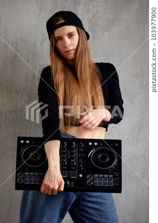 A young pretty long-haired DJ girl in a black top, blue loose jeans and a black baseball cap stands against a gray wall and holds a black DJ mixing console in her hands. Music and fun. A young pretty long-haired DJ girl in a black top, blue loose jeans and a black baseball cap stands against a gray wall and holds a black DJ mixing console in her hands. Music and fun. 103977900