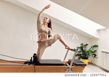 A young girl is doing Pilates on a reformer bed in a bright studio. A slender brunette in a beige bodysuit does exercises to strengthen the spine and back muscles. Healthy lifestyle concept. A young girl is doing Pilates on a reformer bed in a bright studio. A slender brunette in a beige bodysuit does exercises to strengthen the spine and back muscles. Healthy lifestyle concept. 103977952