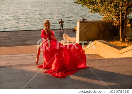 Sunrise red dress. A woman in a long red dress against the backdrop of sunrise, bright golden light of the sun's rays. The concept of femininity, harmony. 103978918