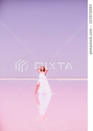 Woman in pink salt lake. She in a white dress and hat enjoys the scenic view of a pink salt lake as she walks along the white, salty shore, creating a lasting memory. 103978983