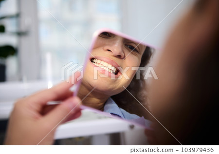 Close-up reflection in the cosmetic mirror of a pretty woman, female patient sitting in dental chair, admiring her beautiful smile and teeth after teeth bleaching procedure in dentistry clinic. 103979436
