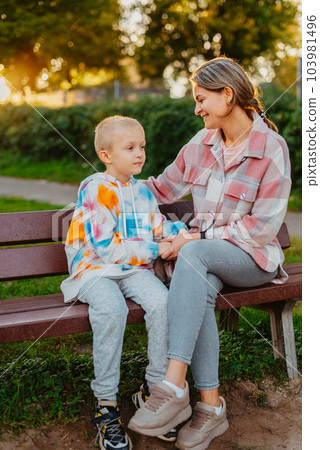 mother and son sit on a park bench in the rays of the setting sun. the concept of a family. Mother's Day. beautiful girl (mother) with a boy (son) in the park in the park are sitting on a bench at 103981496