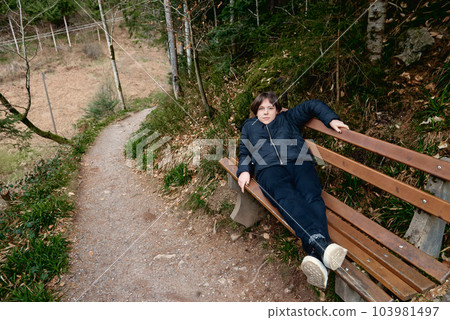 Beautiful young woman in sportswear lying on the wooden bench in the middle of a park without people reading a blue book against a background of green trees. Woman lying on a bench in a park Beautiful young woman in sportswear lying on the wooden bench in the middle of a park without people reading a blue book against a background of green trees. Woman lying on a bench in a park 103981497