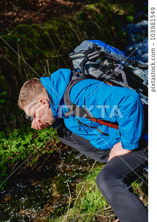Hiker drinking stream water in mountain. Adult man drinking water from the mountain creek. Tourist wearing casual clothes and sunglasses making a sip of mountain river water from the palms of his Hiker drinking stream water in mountain. Adult man drinking water from the mountain creek. Tourist wearing casual clothes and sunglasses making a sip of mountain river water from the palms of his 103981549
