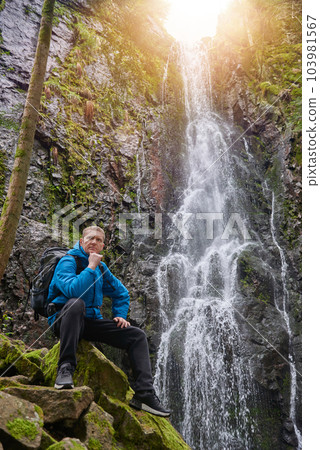 Tourist attraction of Germany - falls of Burgbach Waterfall near Schapbach, Black Forest, Baden-Wurttemberg, Germany. Man hiker in blue jacket standing on stone and looks at flow of falling water 103981567