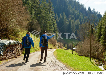 Two hikers are in the mountains in the spring nature outdoor. Hiking with backpacks. Two friends travel together in picturesque places. The man shows the way in Alps. Two hikers are in the mountains in the spring nature outdoor. Hiking with backpacks. Two friends travel together in picturesque places. The man shows the way in Alps. 103981588