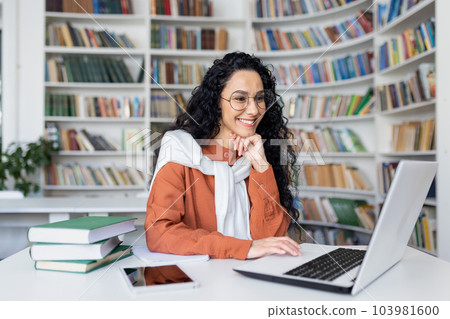 Female student with laptop reading online video course, Latin American woman smiling and satisfied with independent online learning sitting inside university campus in library. Female student with laptop reading online video course, Latin American woman smiling and satisfied with independent online learning sitting inside university campus in library. 103981600