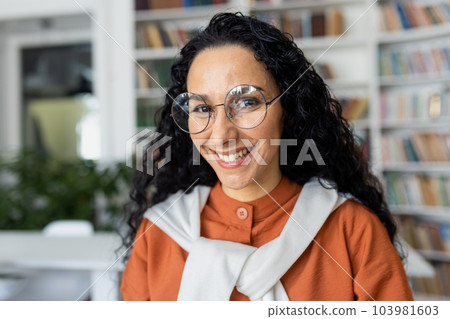 Portrait of young beautiful female student inside university academic library, Hispanic woman with curly hair and glasses close up smiling and looking at camera. Portrait of young beautiful female student inside university academic library, Hispanic woman with curly hair and glasses close up smiling and looking at camera. 103981603
