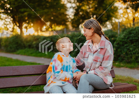 mother and son sit on a park bench in the rays of the setting sun. the concept of a family. Mother's Day. beautiful girl (mother) with a boy (son) in the park in the park are sitting on a bench at mother and son sit on a park bench in the rays of the setting sun. the concept of a family. Mother's Day. beautiful girl (mother) with a boy (son) in the park in the park are sitting on a bench at 103981625