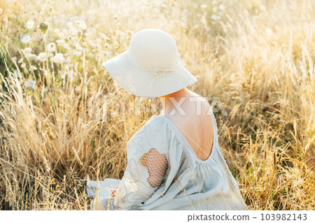 Tender back view woman in light airy dress with open back and straw hat enjoying the moment in a high grass meadow at sunset. summer evening in nature. Calm, harmony. Freckles on body, natural beauty 103982143