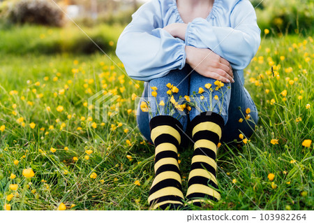 Close up young female wearing jeans and striped black and yellow socks with flowers inside sitting on the green grass of blooming meadow. Concept of bee protection, bloom season, art, creativity 103982264