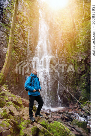 Tourist attraction of Germany - falls of Burgbach Waterfall near Schapbach, Black Forest, Baden-Wurttemberg, Germany. Man hiker in blue jacket standing on stone and looks at flow of falling water Tourist attraction of Germany - falls of Burgbach Waterfall near Schapbach, Black Forest, Baden-Wurttemberg, Germany. Man hiker in blue jacket standing on stone and looks at flow of falling water 103985952