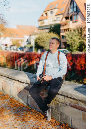 Man Sitting in Old European City And Holding Photo Camera. Contemporary Stylish Blogger And Photographer. Handsome man taking a selfie on a trip in Europe. 103985958
