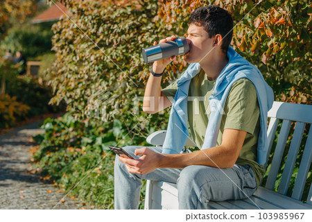 A Teenager Sits On A Bench In The Autumn Park Drinks Coffee From A Thermo Mug And Looks Into A Phone. Portrait Of Handsome Cheerful Guy Sitting On Bench Fresh Air Using Device Browsing Media Smm A Teenager Sits On A Bench In The Autumn Park Drinks Coffee From A Thermo Mug And Looks Into A Phone. Portrait Of Handsome Cheerful Guy Sitting On Bench Fresh Air Using Device Browsing Media Smm 103985967