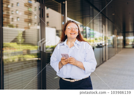 Businesswoman using phone near office. Happy woman in a blue shirt walks on the street with her smartphone. Professional manager working on mobile device. Successful female using modern technology 103986139