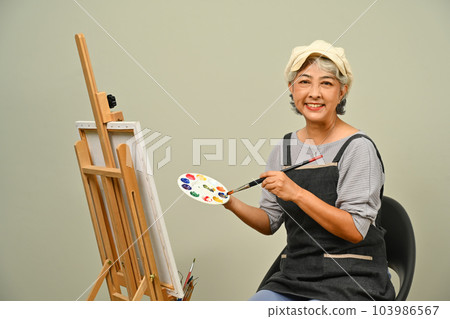 Portrait of beautiful 60s senior woman wearing apron holding brush and palette over gray background Portrait of beautiful 60s senior woman wearing apron holding brush and palette over gray background 103986567