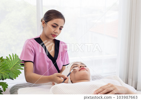 Thai professional  female masseuse therapist in uniform standing using brush brushing essential oil cream on customer face while laying down closed eyes relax on massage bed 103987663