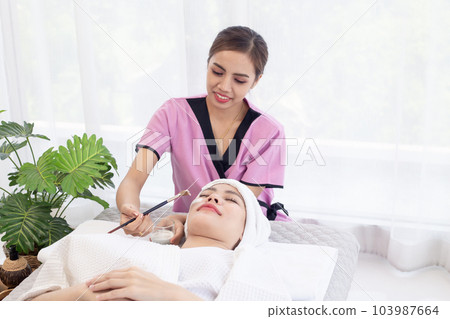 Thai professional female masseuse therapist in uniform standing using brush brushing essential oil cream on customer face while laying down closed eyes relax on massage bed Thai professional female masseuse therapist in uniform standing using brush brushing essential oil cream on customer face while laying down closed eyes relax on massage bed 103987664