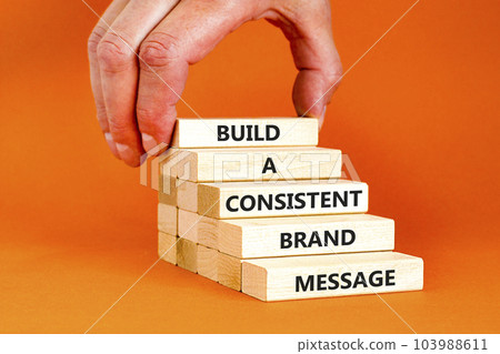 Consistent brand message symbol. Concept words build a consistent brand message on wooden blocks. Businessman hand. Beautiful orange background. Business consistent brand message concept. Copy space. 103988611