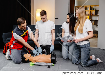 Young man instructor helping to make first aid heart compressions with dummy during the group training indoors Young man instructor helping to make first aid heart compressions with dummy during the group training indoors 103989884