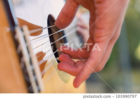Close-up of the musician's playing hands. The guitarist plays the guitar. Professional guitarist plays guitar outdoors. Musician plays a classical guitar in the park. Close-up of the musician's playing hands. The guitarist plays the guitar. Professional guitarist plays guitar outdoors. Musician plays a classical guitar in the park. 103990797