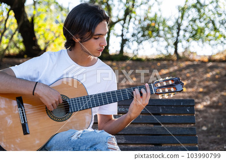 Guitarist plays on guitar outdoors. Handsome guy plays the guitar on the nature sitting on a bench. Young man plays a musical instrument outside house. Musician plays a classical guitar in the Guitarist plays on guitar outdoors. Handsome guy plays the guitar on the nature sitting on a bench. Young man plays a musical instrument outside house. Musician plays a classical guitar in the 103990799