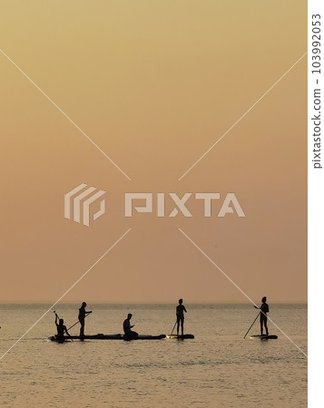 Group of people on stand up paddle board at quiet sea on sunset or sunrise. People on sup board and bright sunset Group of people on stand up paddle board at quiet sea on sunset or sunrise. People on sup board and bright sunset 103992053
