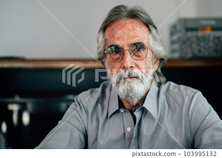 Portrait of senior man with grey hair and beard sitting on sofa at home Portrait of senior man with grey hair and beard sitting on sofa at home 103995132